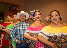Five folklórico dancers pose in a diagonal line indoors, wearing colorful traditional outfits with floral skirts, off-the-shoulder blouses, flower headpieces, and gold earrings. One dancer in the center wears a white cowboy hat, plaid shirt, jeans, and belt buckle.