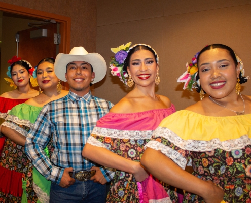 Five folklórico dancers pose in a diagonal line indoors, wearing colorful traditional outfits with floral skirts, off-the-shoulder blouses, flower headpieces, and gold earrings. One dancer in the center wears a white cowboy hat, plaid shirt, jeans, and belt buckle.