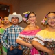 Five folklórico dancers pose in a diagonal line indoors, wearing colorful traditional outfits with floral skirts, off-the-shoulder blouses, flower headpieces, and gold earrings. One dancer in the center wears a white cowboy hat, plaid shirt, jeans, and belt buckle.