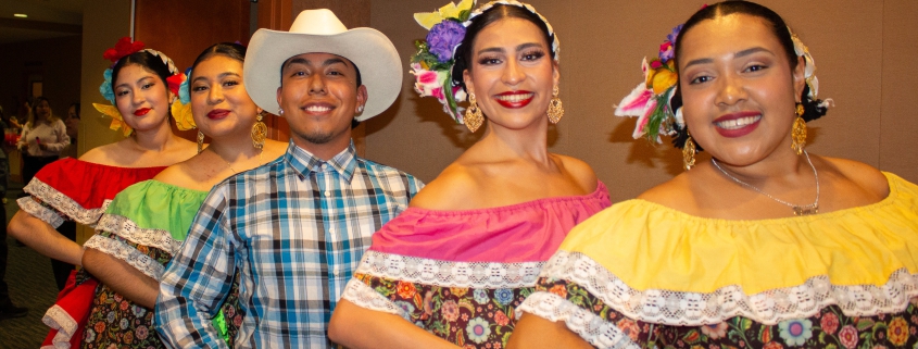 Five folklórico dancers pose in a diagonal line indoors, wearing colorful traditional outfits with floral skirts, off-the-shoulder blouses, flower headpieces, and gold earrings. One dancer in the center wears a white cowboy hat, plaid shirt, jeans, and belt buckle.